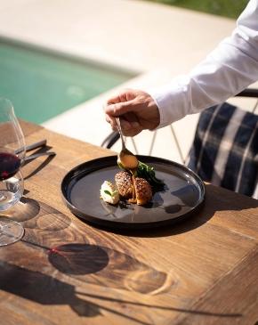 Chef serve piatto gourmet con salsa, accanto a bicchiere di vino rosso.
