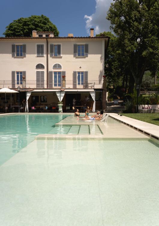 Villa con piscina, persone rilassate al sole, atmosfera tranquilla e alberi circostanti.