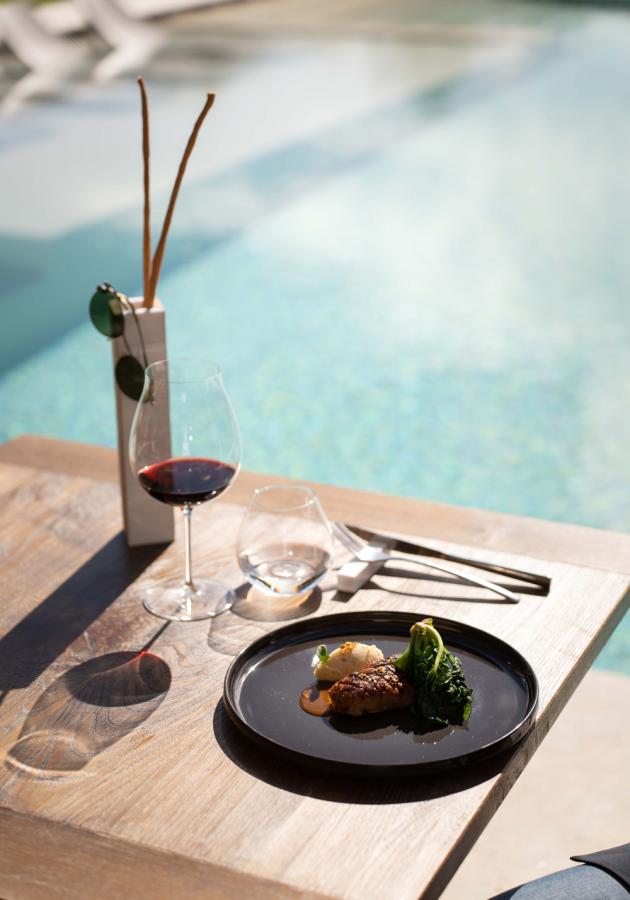 Pasto elegante a bordo piscina con vino rosso e piatto gourmet su tavolo di legno.