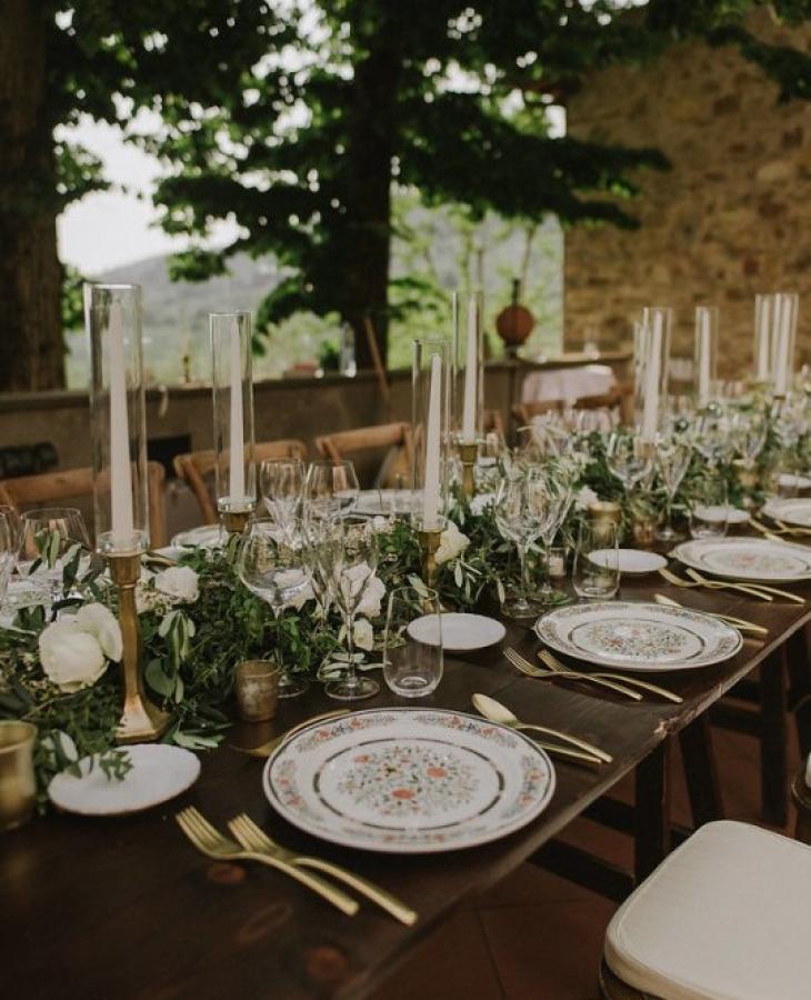 Tavola elegante all'aperto con candele, piatti decorati e centrotavola verde.