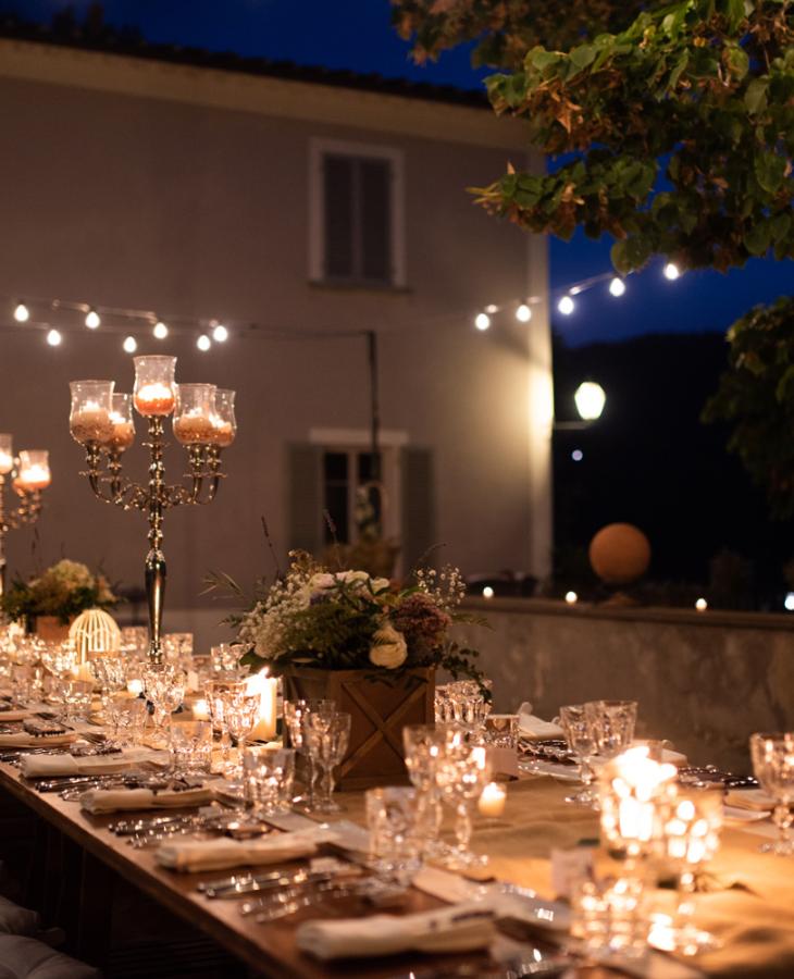 Tavola elegante all'aperto, illuminata da candele e luci, atmosfera serale accogliente.