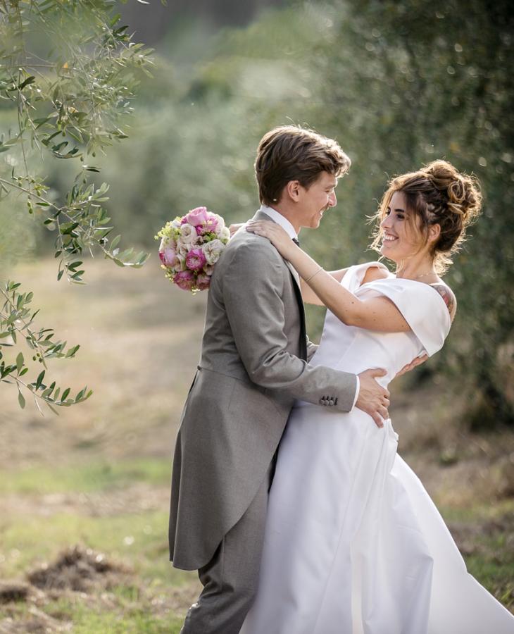 Coppia di sposi felici in un uliveto, con abiti eleganti e bouquet.